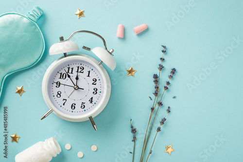 Top view photo of white alarm clock lavender blue satin sleeping mask pink earplugs open bottle with pills and golden stars on isolated pastel blue background