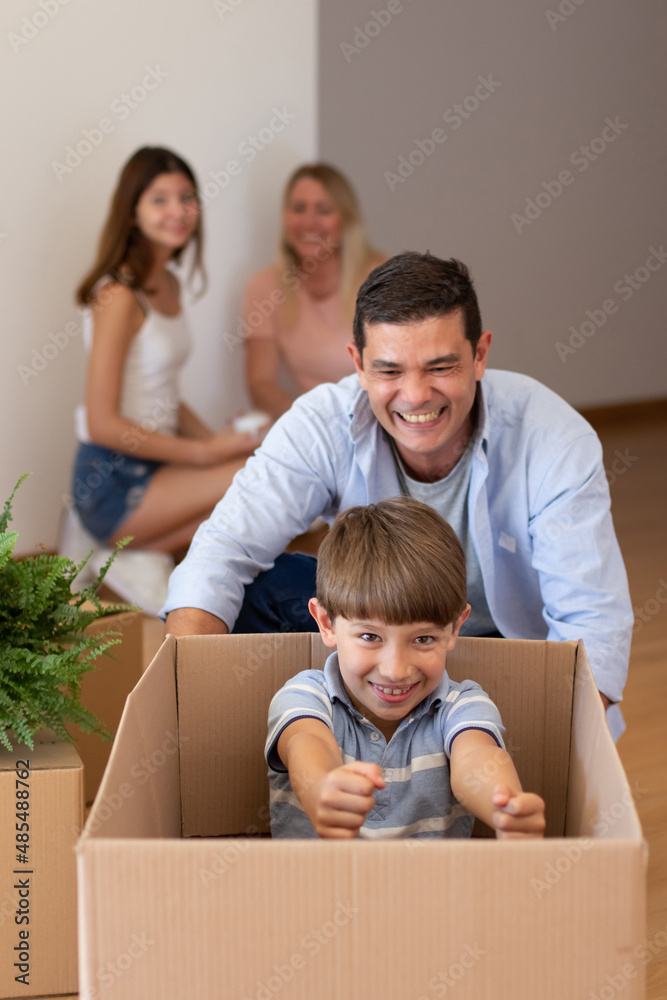 Merry mid adult father playing with son. Schoolboy with dark hair sitting in big cardboard box. Man riding him in it, imitating car. Real estate, purchase, family concept Merry mid adult father playing with son. Schoolboy with dark hair sitting in big cardboard box. Man riding him in it, imitating car. Real estate, purchase, family concept