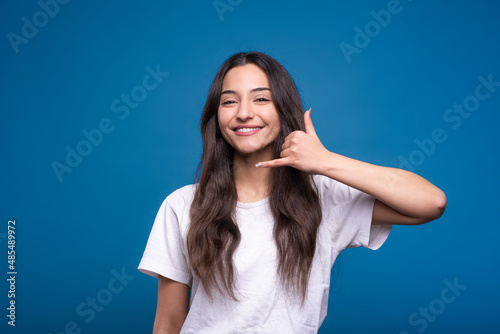 Fotografie Attractive caucasian or arab brunette girl in a white t-shirt showing a call gesture on a mobile phone isolated in on a blue studio background