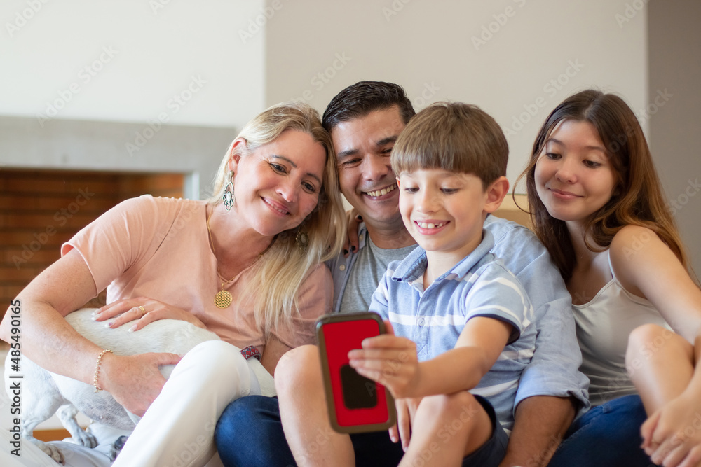 Merry family taking selfie in new house. Mid adult parents, teenage girl and little boy together, looking at mobile phone camera smiling. Hugging. Social media, purchase, family concept