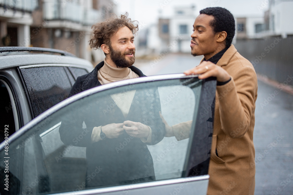 Two men flirting or having a close conversation while opening car door on the street. Concept of homosexual relations or close male friendship. Caucasian and hispanic man wearing coats Two men flirting or having a close conversation while opening car door on the street. Concept of homosexual relations or close male friendship. Caucasian and hispanic man wearing coats