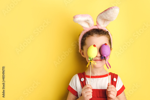 Portrait of funny little girl covers eyes with two Easter eggs, wears bunny ears, posing isolated over yellow studio background with copy space for advertisement. Spring holiday and Easter concept