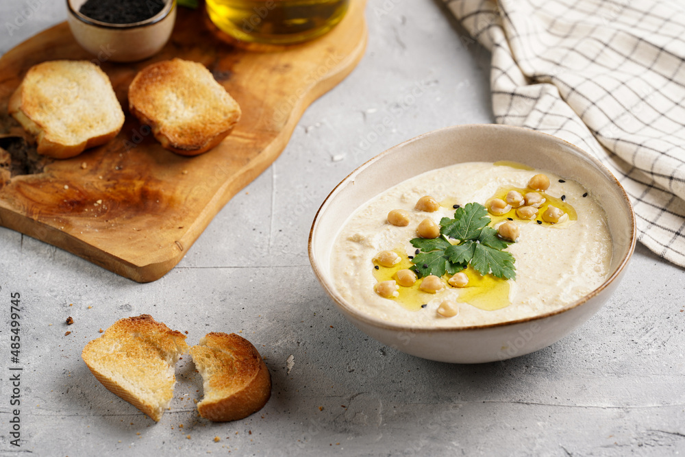 Hummus dip with chickpea and parsley in ceramic bowl on concrete surface with toasted baguette slices and olive oil on wooden board Hummus dip with chickpea and parsley in ceramic bowl on concrete surface with toasted baguette slices and olive oil on wooden board