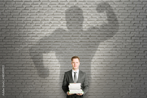 Abstract image of attractive european businessman with books and strong muscle shadow on brick wall background. Confidence and strength concept. © Who is Danny