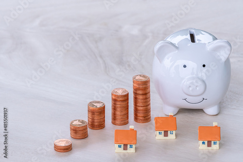 Piggy bank with stacks of coins are placed next to each other in a growing direction and many houses are placed on wooden tables, Saving money buy a house in the future and how to buy a house concept.