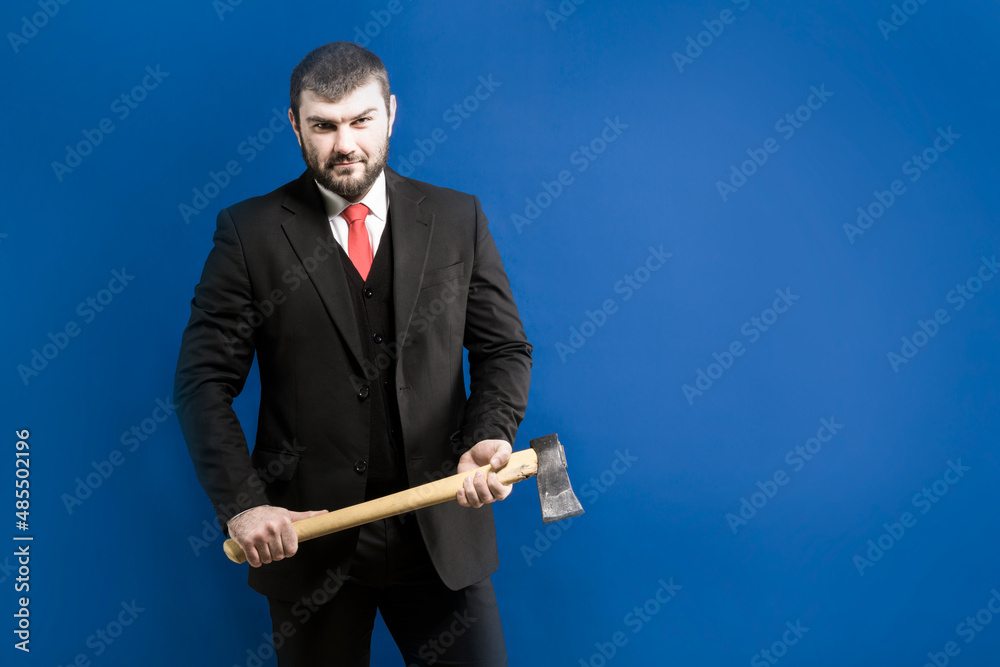Revenge. Mafia. Businessman. Maniac. Dangerous guy. Serial killer. A man with a beard in a suit and a red tie holds an ax in his hands and looks confidently ahead on a blue background Revenge. Mafia. Businessman. Maniac. Dangerous guy. Serial killer. A man with a beard in a suit and a red tie holds an ax in his hands and looks confidently ahead on a blue background