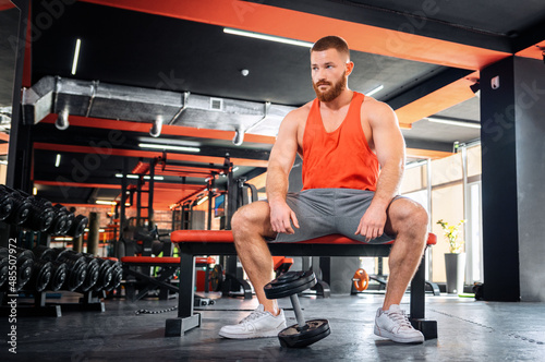 A young bearded athletic man is sitting on a sports bench. The dumbbell lies near the athlete's feet. Bodybuilding and fitness concept