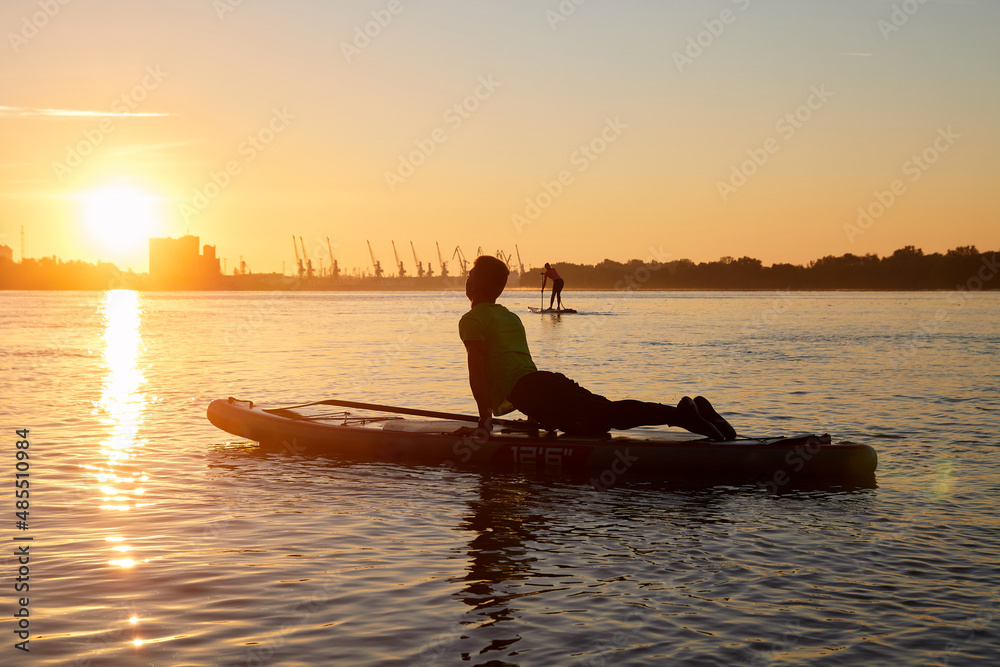Healthy and fit man standing in yoga asana on paddle board at Danube river. Silhouette of young guy doing morning exercises on nature.