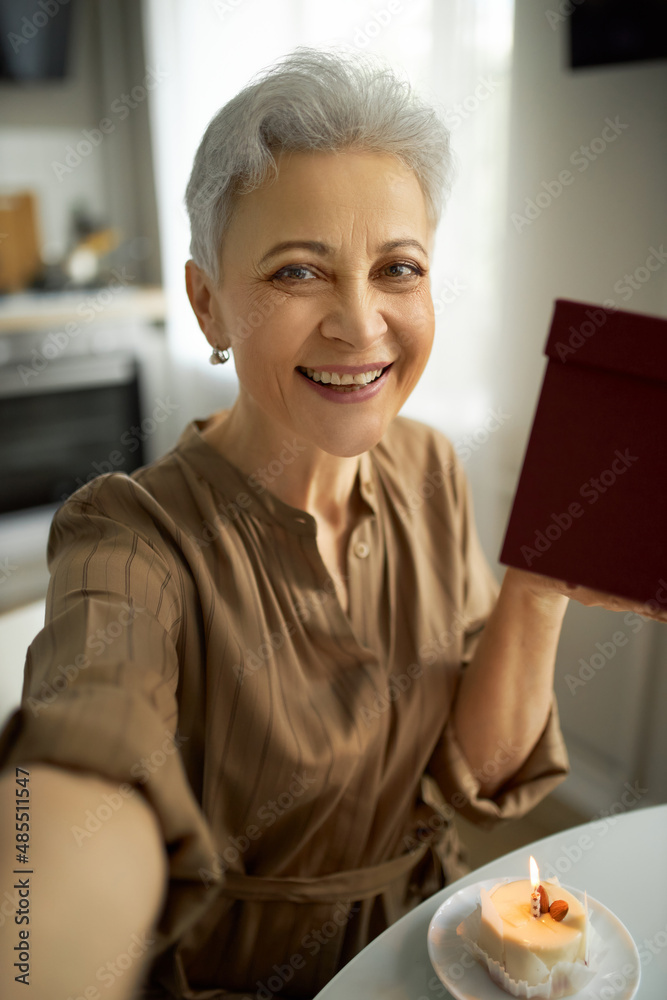 Closeup of pretty mature Caucasian female with grey hair sitting at kitchen table with present box, doing selfie, smiling widely. Human emotions and feelings. Technology and everyday life Closeup of pretty mature Caucasian female with grey hair sitting at kitchen table with present box, doing selfie, smiling widely. Human emotions and feelings. Technology and everyday life