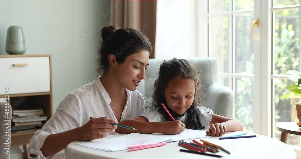Indian mother cute little daughter drawing in sketchbook with colored pencils, babysitter and preschool girl enjoy pastime, talk sit at table in cozy living room. Creative hobby, development concept Indian mother cute little daughter drawing in sketchbook with colored pencils, babysitter and preschool girl enjoy pastime, talk sit at table in cozy living room. Creative hobby, development concept