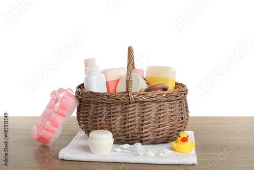Wicker basket full of different baby cosmetic products, bathing accessories and toys on wooden table against white background