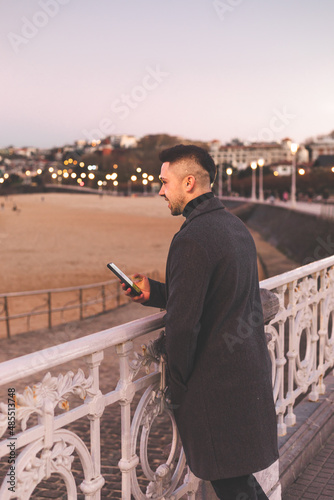 Young caucasian man using a smartphone at Donostia-San Sebastian sea walk during sunset time.