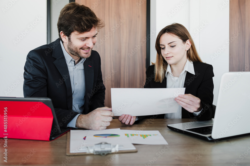 Businesswoman discussing report with colleague sitting at desk in office Businesswoman discussing report with colleague sitting at desk in office
