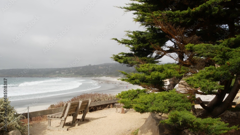Empty wooden bench, rest on trail path, walkway or footpath. Carmel beach, Monterey ocean shore, California coast USA. Sea waves crashing. Waterfront beachfront pine cypress trees. Pathway or footway.