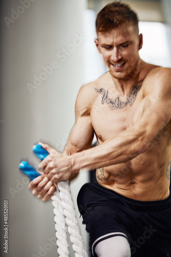 Muscular powerful aggressive man training with rope in functional training fitness gym. Motion blur