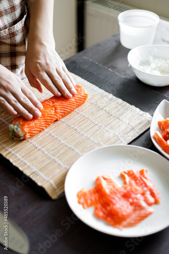 Young woman wrapping up sushi roll ingredients on bamboo mat. Process of making sushi at home