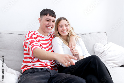 Fototapeta Naklejka Na Ścianę i Meble -  Boyfriend and girlfriend watching TV laughing. Boy pointing to the screen. Young couple at home. Heterosexual 18-20 years old couple.