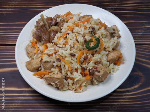 Traditional national dish pilaf. Garlic, carrots, rice, meat with seasonings and sauces in a plate on a wooden table, flat layout. Delicious Uzbek hot food plov with chicken, cereals and vegetables