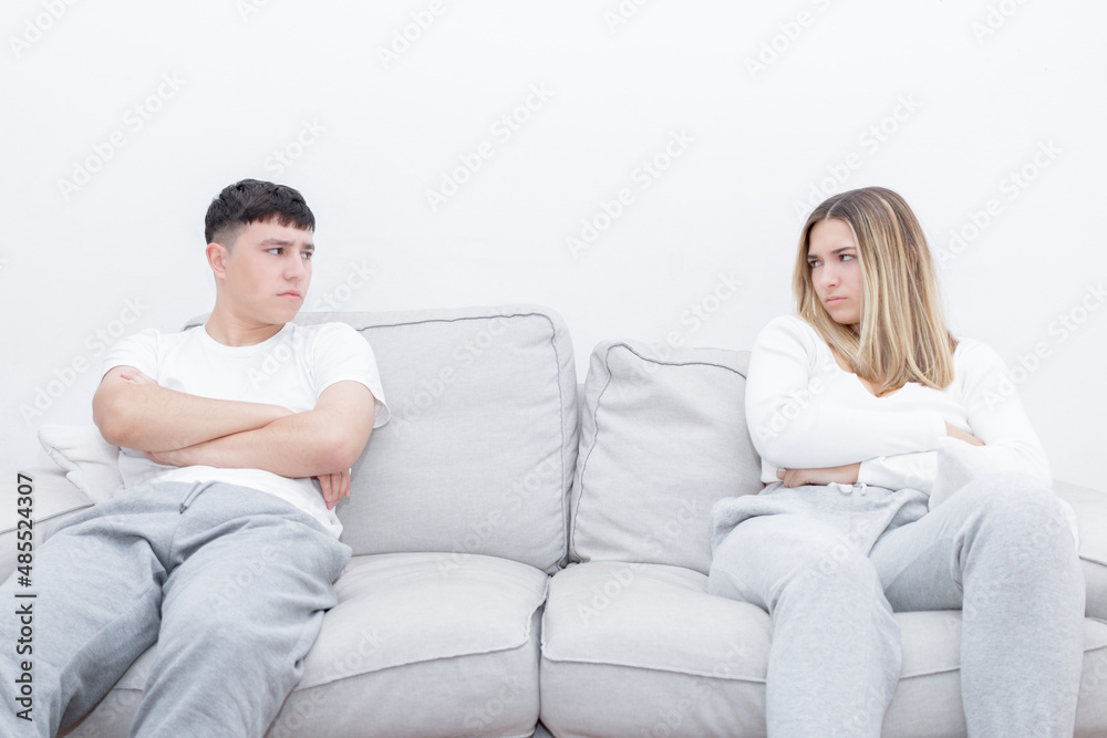 Angry couple sitting separated in the sofa looking at each other. Mad couple after a relationship conflict. Young couple at home. Heterosexual 18-20 years old couple.