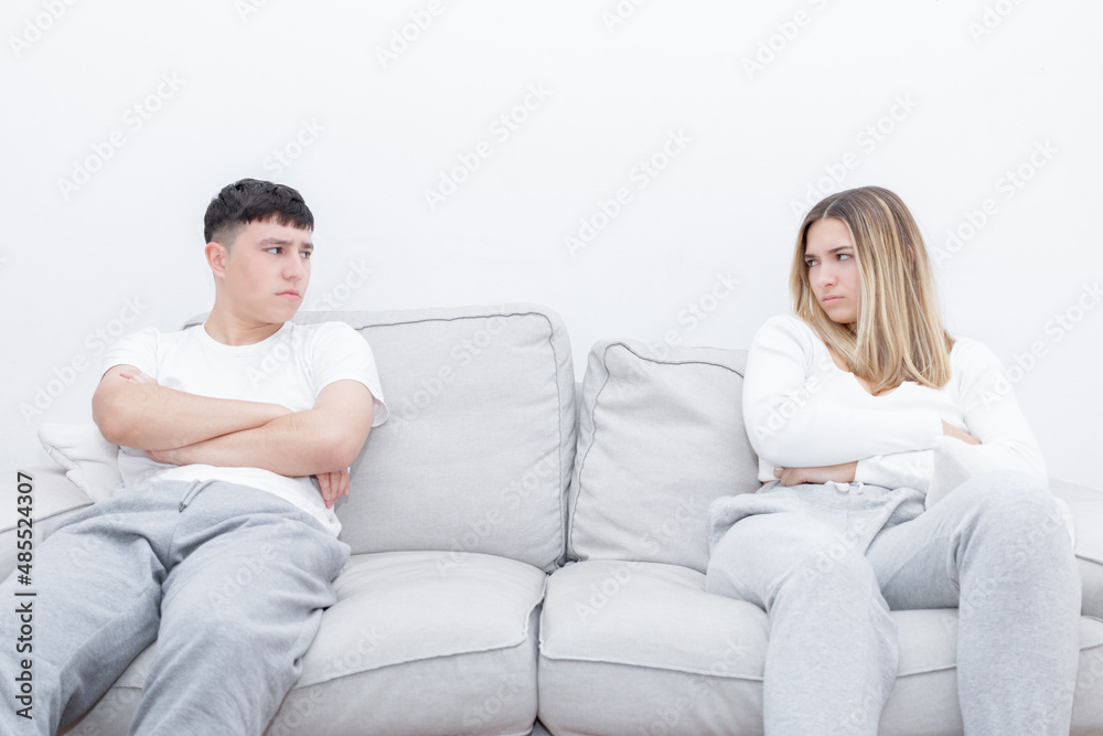 Angry couple sitting separated in the sofa looking at each other. Mad couple after a relationship conflict. Young couple at home. Heterosexual 18-20 years old couple. Angry couple sitting separated in the sofa looking at each other. Mad couple after a relationship conflict. Young couple at home. Heterosexual 18-20 years old couple.
