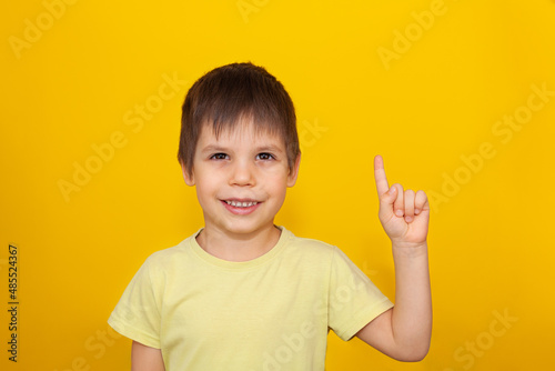 Portrait of smiling little boy with finger pointed up. Kid isolated on yellow blackboard. Copy space.