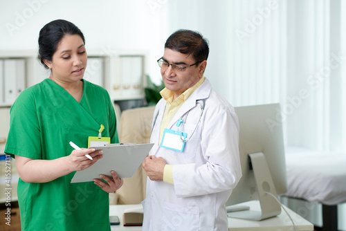 Focused mature male doctor and female nurse looking at screen disc of tablet computer and discussing anamnesis of patient