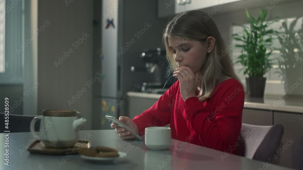 Beautiful teenage girl sitting in the kitchen at the table with a cup of tea and looking at the phone. Modern baby breakfast with phone.