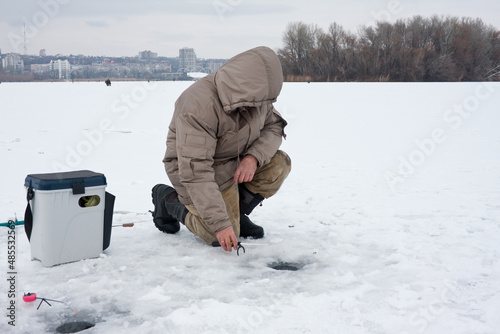 Man on winter fishing. The fisherman is on one knee and sets up a fishing rod. A man fishes on an ice-bound river. Winter cityscape. photo
