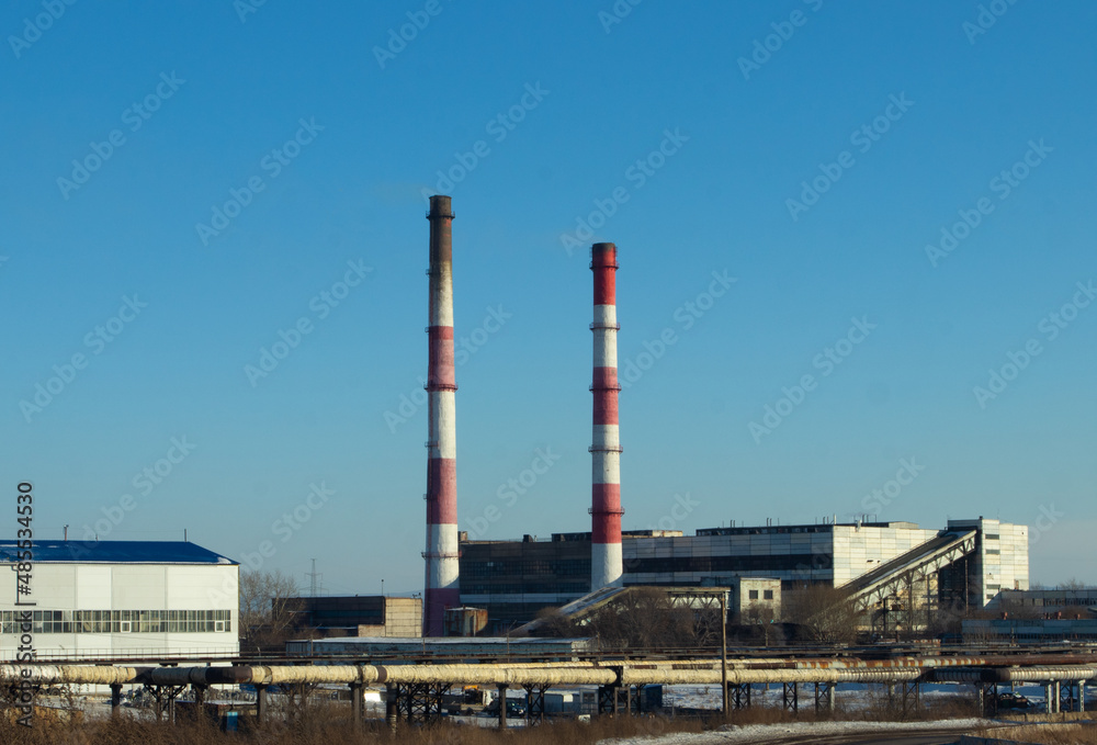 High red and white striped tube lets out white smoke into clear blue sky. Small cloud above pipe on background of electric pole and power wires. Thermal power plant close-up with copy space.