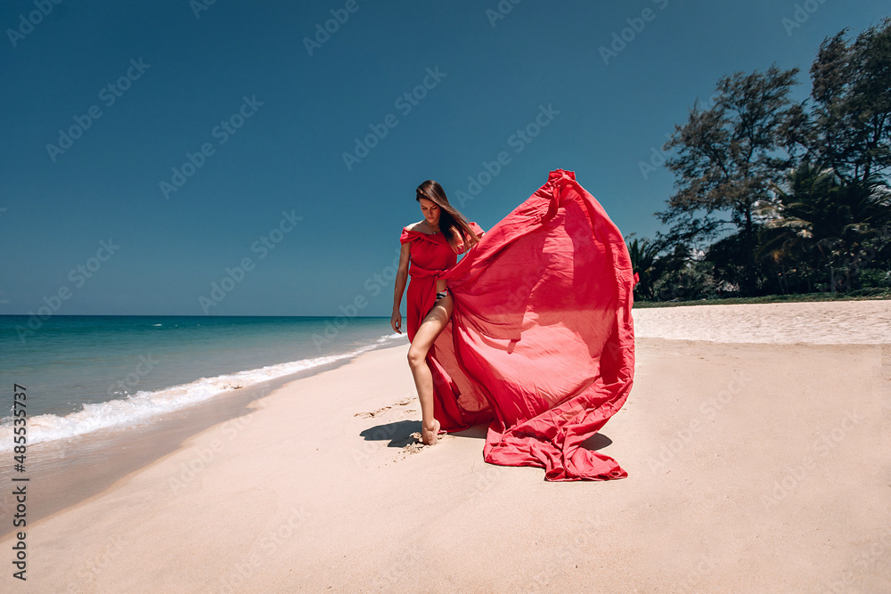 Stunning girl in the exotic sandy beach by the sea. The lady wears light red dress that flows in the wind; fashion concept. Stunning girl in the exotic sandy beach by the sea. The lady wears light red dress that flows in the wind; fashion concept.