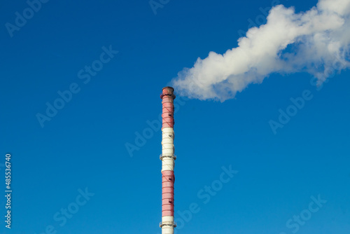 High red and white striped tube lets out white smoke into clear blue sky. Small cloud above pipe on background of electric pole and power wires. Thermal power plant close-up with copy space.