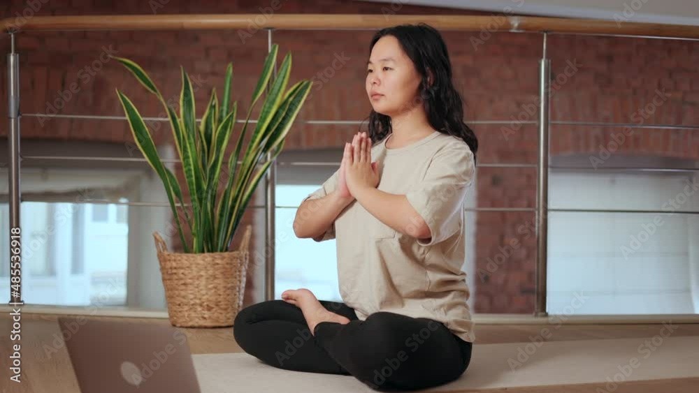 Young woman meditating and doing yoga while sitting on floor in home room spbi. Close-up view of Asian female sits in lotus position and joins her hands in mudra, meditates calmly and does oriental Young woman meditating and doing yoga while sitting on floor in home room spbi. Close-up view of Asian female sits in lotus position and joins her hands in mudra, meditates calmly and does oriental