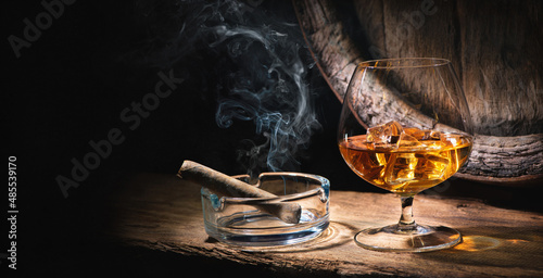 Glass of whiskey with smoking cigar and ice cubes in front of old barrel