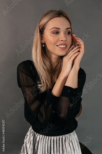 Wonderful pretty european girl with blond hair wearing black blouse and silver skirt gently touching her face and looking away with wonderful smile over grey background