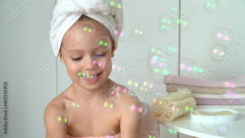 Portrait of little girl with wrapped towel on head laughs and plays soap bubbles in bathroom. Happy kid after bathing. Baby care. Soap and shampoo concept photo