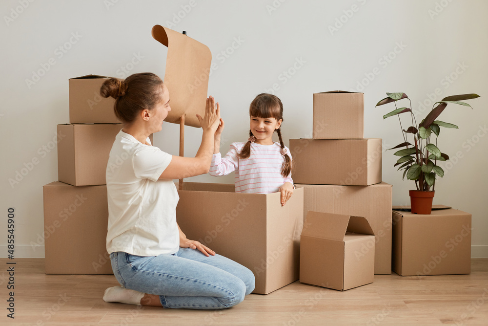 Indoor shot of attractive woman playing with her daughter, making ship with flag during relocation and playing sailor, resting while unpacking personal pile, giving five each other.