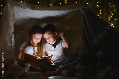 Little kids involving in reading amazing book. They lying in nice toy tent in playroom. Boy holding flashlight in hand