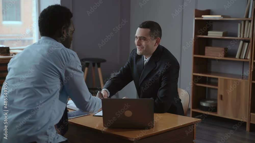 Two businessmen shaking hands and talking while sitting at table with laptop in company spbas. Close-up view of young multi-ethnic men shake hands and look with smiles, talk and sit at desk with
