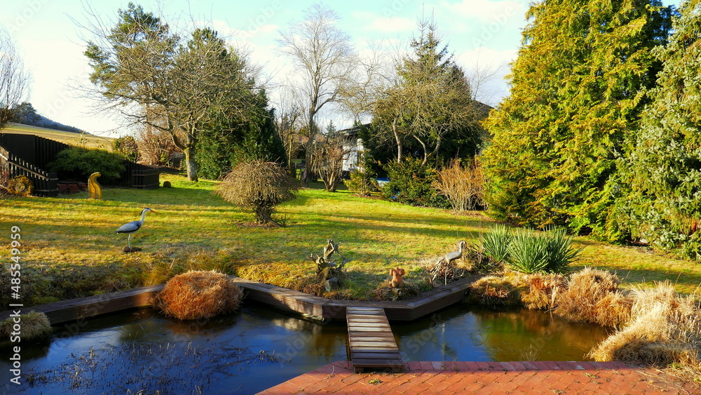 malerischer sonniger Gartenteich im Winter an roter Terrasse mit Holzbrücke, Wiese und Bäumen mit Raureif malerischer sonniger Gartenteich im Winter an roter Terrasse mit Holzbrücke, Wiese und Bäumen mit Raureif