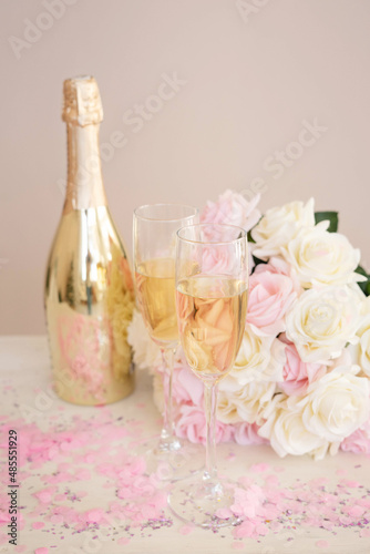 Two glasses, a golden bottle of champagne, a bouquet of roses and pink confetti on the table. Romantic date on Valentine's Day.