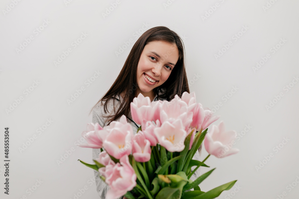 Wonderful happy woman with dark hair and lovely happy smile posing at camera with tender pink flowers over isolated background. Happy woman is celebrating Woman's Day Wonderful happy woman with dark hair and lovely happy smile posing at camera with tender pink flowers over isolated background. Happy woman is celebrating Woman's Day