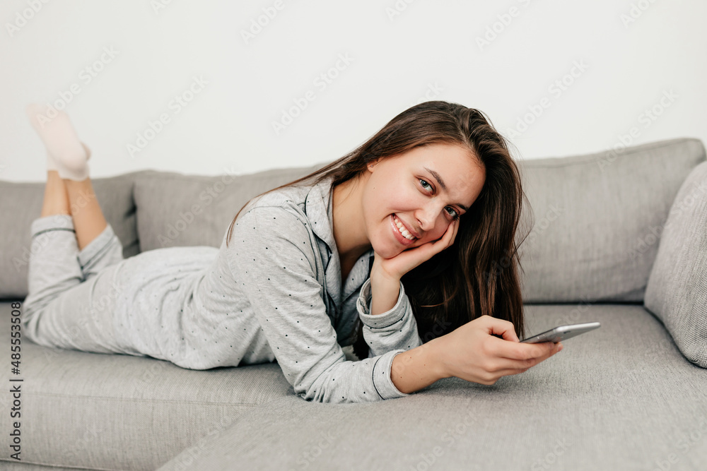 lovely pretty girl with long dark hair is wearing home suit lying on sofa and smiling at camera with smartphone. Indoor portrait of happy girl is relaxing at home lovely pretty girl with long dark hair is wearing home suit lying on sofa and smiling at camera with smartphone. Indoor portrait of happy girl is relaxing at home