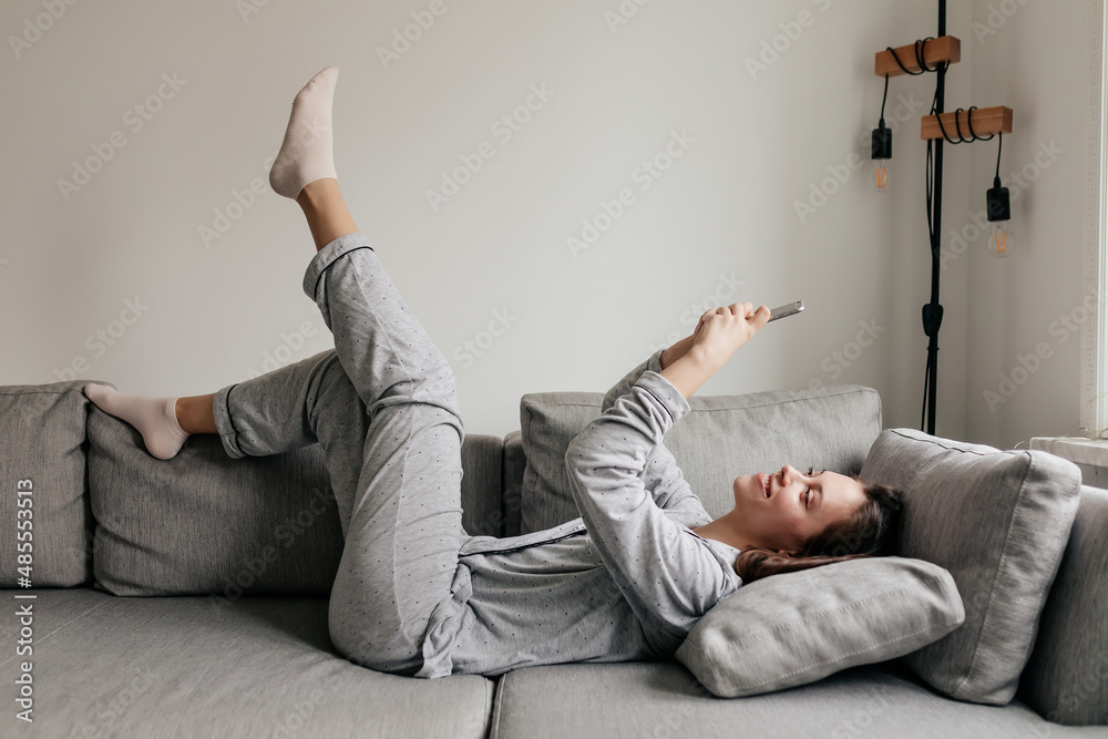 Full-lenght portrait of happy excited girl is lying on sofa in home suit and using smartphone with smile. Excited lovely girl is holding up leg and spend free time at home