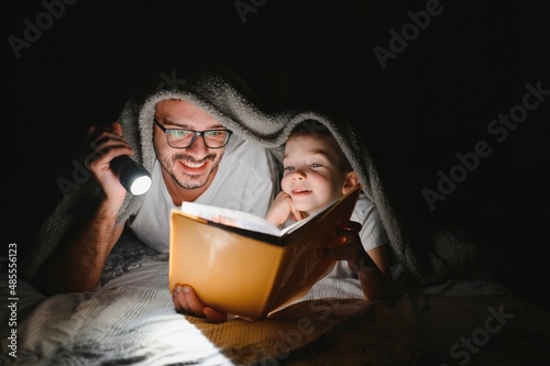 Father and son with flashlight reading book under blanket at home photo
