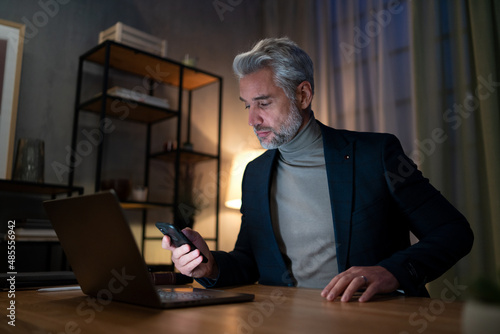 Mature businessman working on laptop at desk indoors in office at night.