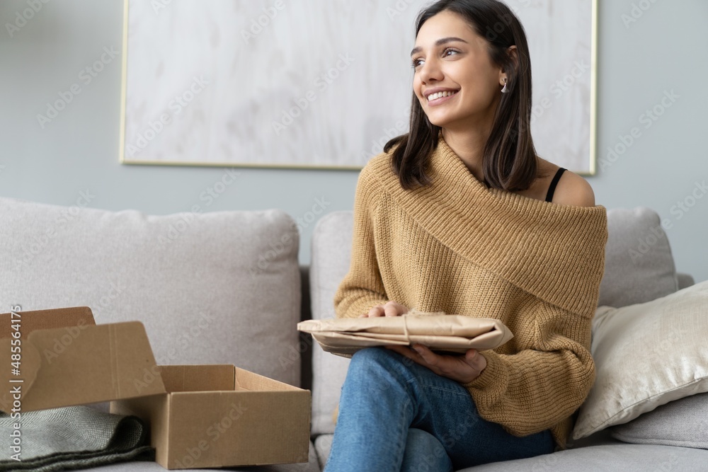 Smiling happy young woman sitting on couch, unpacking carton box parcel online order from international internet store