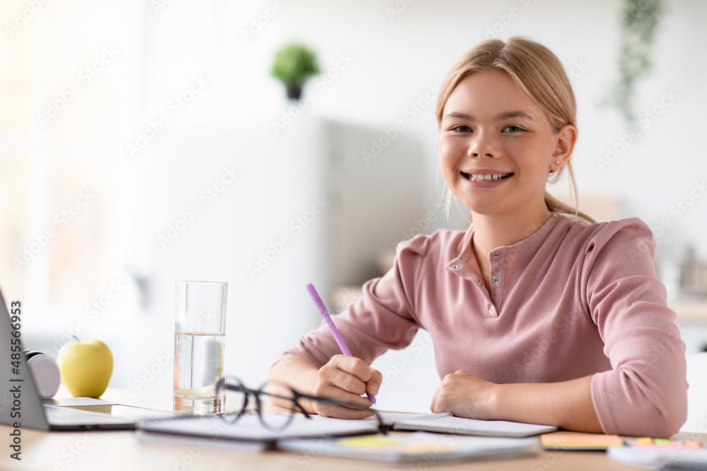 Smiling european adolescent girl blonde pupil study at home at table with laptop in light kitchen interior Smiling european adolescent girl blonde pupil study at home at table with laptop in light kitchen interior