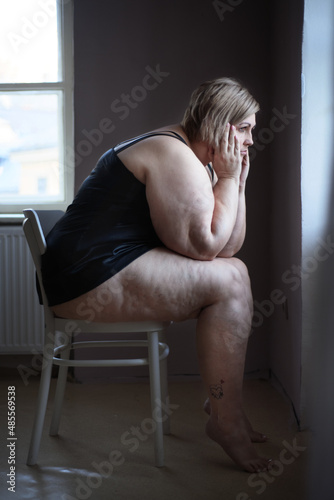 Depressed lonely fat woman sitting and looking through the window at home.
