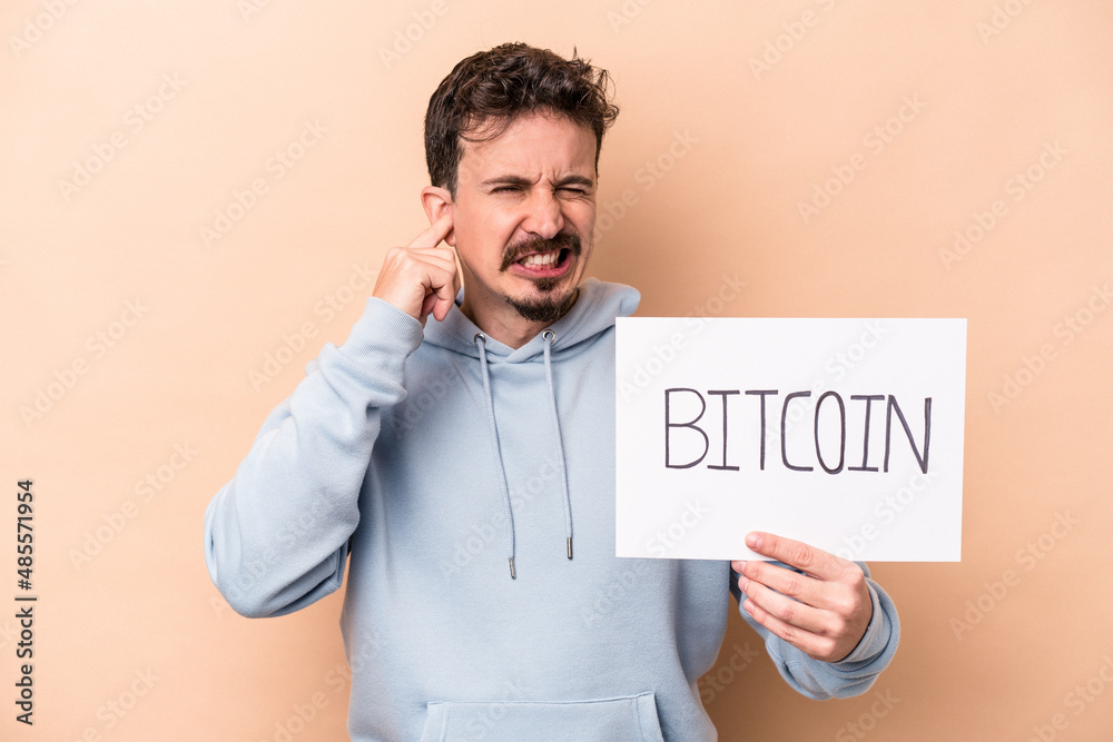 Young caucasian man holding a bitcoin placard isolated on beige background covering ears with hands. Young caucasian man holding a bitcoin placard isolated on beige background covering ears with hands.