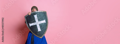 Portrait of scary man, medieval warrior or knight in armor hiding himself behind a shield isolated over pink studio background. Flyer